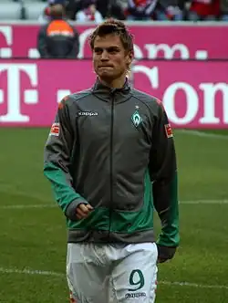A photograph of a blonde man wearing a grey and green training jacket and white shorts, the man is seen on a football pitch in the process of warming up.