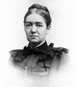 An older Caucasian female with parted dark hair swept back into a bun. She is wearing a dark high collared shirt with ruffles and facing the camera unsmiling.
