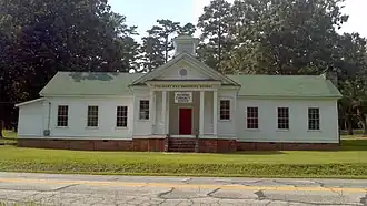 A white one-room schoolhouse on a small road, with a white exterior, a green roof, and a red front door.
