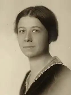 Black and white photograph of the bust of a young white woman before a white background with dark hair parted in the middle and tied up in the back, a serious countenance with eyes on the viewer, and a dark jacket over a white blouse.