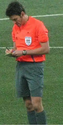 A man wearing a bright orange shirt and grey shorts, writing in a notebook. He has dark hair.