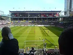 A soccer match in a crowded multilevel stadium on a sunny day. There are Swedish flags on the top of the back wall, and the back awning reads “VI ÄLSKAR NÄR SVERIGE VINNER”.