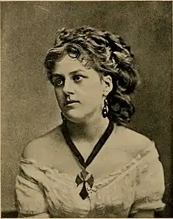 A white woman with light hair in an elaborate curled coiffure, wearing an off-the-shoulder light dress and a large cross-shaped pendant on a dark ribbon