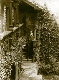 A man with a greying beard wearing a hat standing on the steps of a castle, slightly leaning on the railing whilst vines frame him.