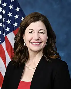 Official House portrait of McDonald Rivet smiling in front of the U.S. flag, wearing a black jacket, red shirt, and pearl necklace.