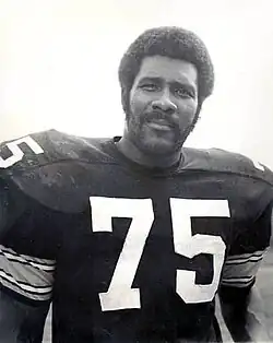 A man with an Afro and a beard in a black football uniform with 75 written in white