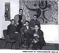 Black and white photo of five men at an art gallery. One stands, two sit in chairs, one sits on the arm of a chair, and one kneels on the floor.