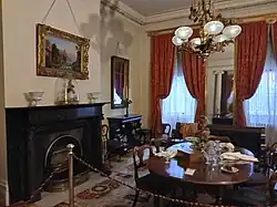 The rear parlor, which has a dining table and a piano. There is a chandelier above, and two windows with curtains behind.