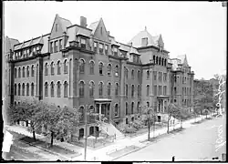 An elevated view of a five-story building on a street corner that takes up a long portion of a block. The building has multiple entrances leading to the sidewalk on the right. Trees line the street rising to the height of about two stories.