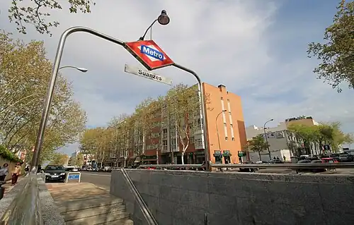 Photograph of a street from the pavement. From a height of about 1 meter we see stairs leading downwards covered by a metallic arch with a rhombus with the Madrid Metro logo and a rectangular sign reading "Suanzes". On the other side of the street, a five-floor building. There are tall trees on both sides of the street.