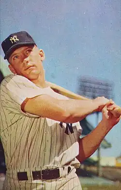 "A young man in New York Yankees pinstripes and cap looks ahead whilst swinging a baseball bat."