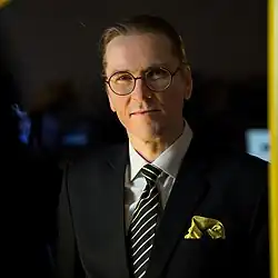 Portrait of Mikko Hyppönen, a Finnish man in his early fifties. He is tall and white, with a high forehead, large ears, and bushy eyebrows that show some gray. He wears round glasses and has light brown, almost reddish hair tied back in his signature ponytail (though the ponytail itself is not visible in the frame). He is dressed formally in a dark jacket, white dress shirt, a tie with wide dark stripes and alternating narrow white and pale yellow stripes, and a golden-yellow pocket square. He looks directly at the camera with a serious expression. Behind him is a blurred nighttime skyline with scattered lights and a golden-yellow vertical bar along the right edge of the image.