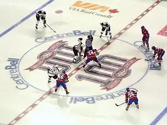 Several ice hockey players in red uniforms line up around the near half of a large circle on the ice, opposed on the other side by several in white uniforms. A player from each team stands at the centre of the circle, joined by a referee wearing a vertically striped black and white uniform.