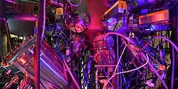 Close-up of a complex stainless-steel scientific instrument with multiple vacuum chambers, circular flanges, cables, and labeled ports, illuminated by vivid purple and red lighting, suggesting a high-precision laboratory system such as molecular beam epitaxy or vacuum deposition equipment.