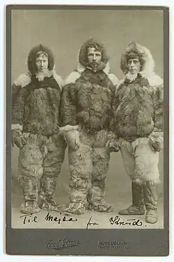 Three men in heavy fur parkas, trousers, and boots stand side by side in a posed studio portrait, wearing cold-weather clothing suitable for Arctic travel, against a plain background.