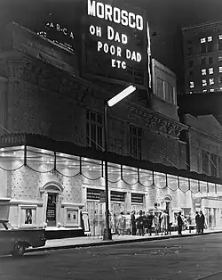 The Morosco Theater as seen from across the street at night. Under the marquee are a group of people. There is a sign displaying the theater's name and the name of the play "Oh Dad, Poor Dad, Etc."