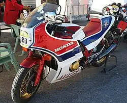 Red white and blue motorcycle with gold wheels in a race paddock against a background of commercial vehicles and camping equipment