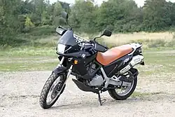 A black BMW F650 fitted with an orange seat, parked on an area of open ground