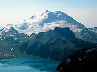 A prominent, glaciated mountain rising over a flat-topped, rocky mountain and a turquoise-coloured alpine lake in the foreground.