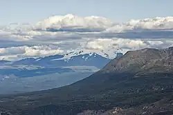 A glaciated, flat-topped, gently-sloping mountain shadowed by clouds with a much lower barren mountain in the right foreground