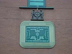 A stone tablet, inscribed with the image of a football pitch and several names. It is surrounded by a stone border in the shape of a football stadium. Above the tablet is a wooden carving of two men holding a large wreath.