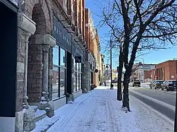 A snow-covered sidewalk runs alongside a long, reddish-brown stone building with rough masonry walls, tall dark-framed storefront windows, and carved stone arches. A bare tree stands near the curb, and the winter sky is clear and blue.