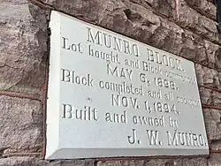 A light grey stone plaque with raised lettering is set into a wall of darker, rough-cut stone. The surface appears smooth and engraved, contrasting with the uneven texture of the surrounding masonry.