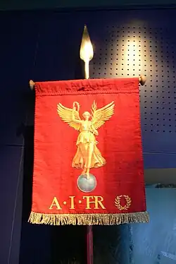 An red upright banner suspended from a pole, on which is depicted a winged figure of victory, holding a laurel wreath, surmounting a globe. Below are the letters A. I. T.R.