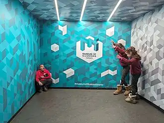 Three people standing in an exhibit in the Toronto Museum of Illusions. Two of the child attendees are perceived to be larger than the third adult attendee.