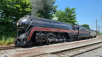 A black streamlined steam locomotive with a 4-8-4 wheel arrangement (four leading wheels, eight driving wheels, and four trailing wheels) and its tender, pulling an observation car with the trees and overhead lines in the background