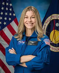 An woman in a blue flight suit is sitting in front of US & NASA flags; she is facing the camera with crossed arms and is smiling.