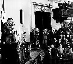 To the left, a man in a suit and tie is standing at a podium in front of the Egyptian flag. To the right, a crowd of people watch, most of whom are men in suits. Some of the men are holding a camera.
