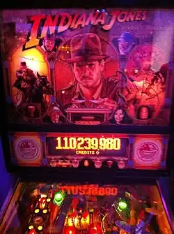 The image contains a backbox of a Indiana Jones themed pinball machine. The backbox shows a image of Indiana Jones and below it with a screen telling the player they got 110,239,980 points.