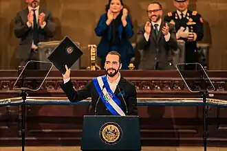 Nayib Bukele standing at a podium and holding up a legislative proposal in his right hand