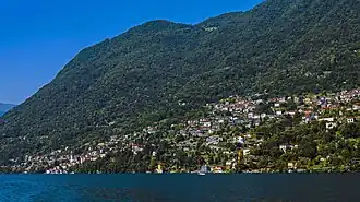 A large settlement consisting mostly of light-colored structures with orange roofs rising up the lower slopes of forested mountains seen from a body of water under a blue sky