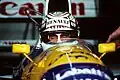 Nigel Mansell wearing a crash helmet with sponsors logos and sitting inside a blue, yellow and white racing car