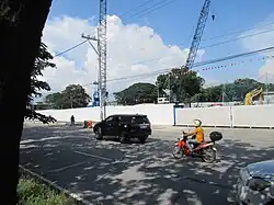 A construction site walled off by white fences. Cranes and an excavator can be seen in the background.