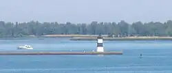 A pier with a square, black and white-striped lighthouse at the end. A small motorboat is paralleling the pier, heading away from the lighthouse.