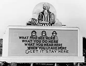 Uncle Sam has removed his hat and is rolling up his sleeves. On the wall in front of him are three monkeys and the slogan: What you see here/ What you do here/ What you hear here/ When you leave here/ Let it stay here.