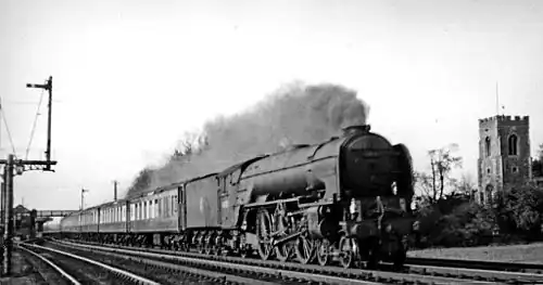 A steam train photographed in black and white at speed from a person by the tracks, clearly lower down. The train is long and the locomotive is clearly producing a great deal of smoke. To its right in the background is a medieval church. To its left in the foreground is a semaphore signal mounted on a gantry.