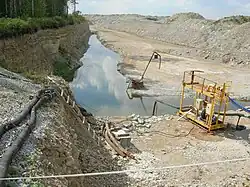 A colour photograph of the Põhja-Kiviõli oil shale mine near Kohtla-Järve, dated to 2007. Machinery and supporting devices are visible in the lower half and a stream of water traverses the centre of the photo. A cliff on the left is topped with trees; the low hills on the right are bare.
