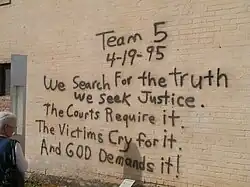A woman, at the left of the image, is reading a black spray paint message written on a brick wall. The message reads "Team 5 4–19–95 We Search For the truth We Seek Justice. The Courts Require it. The Victims Cry for it. And God Demands it!"