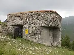 Low concrete bunker with a metal door and firing embrasures, set in a grassy alpine landscape.