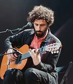 close-up of a seated José González wearing a heavier dark shirt over a light pink one, with dark pants, playing an acoustic guitar in front of a microphone onstage