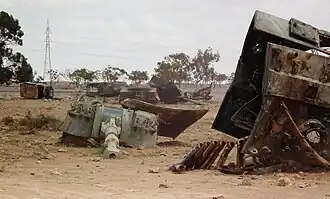 Pieces of a destroyed tank, notably the gun turret, lie on a sandy landscape.
