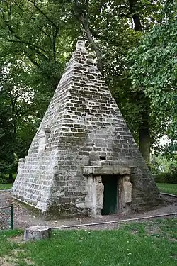 Small stone pyramid with doorway framed by carved figures, surrounded by trees and grass