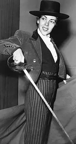 Black and white photograph of a woman wearing a bullfighter's costume, she is holding a sword in one hand and a cape in the other.