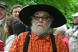 An older man with a white beard and glasses wears a wide-brimmed black hat, red plaid shirt, and suspenders while standing in a crowd outdoors