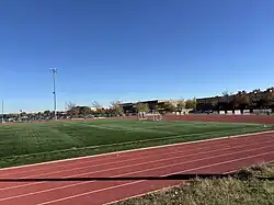 This photograph looks east across the athletics complex at Peak to Peak Charter School in Lafayette, Colorado, showing the 8-lane rubber track encircling a synthetic-turf soccer pitch, portable soccer goals at midfield, a tall light standard at left, and the school buildings and spectator area beyond the perimeter fence.