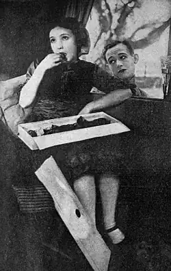 Young woman eating chocolates from a box while a young man gazes at her through a window